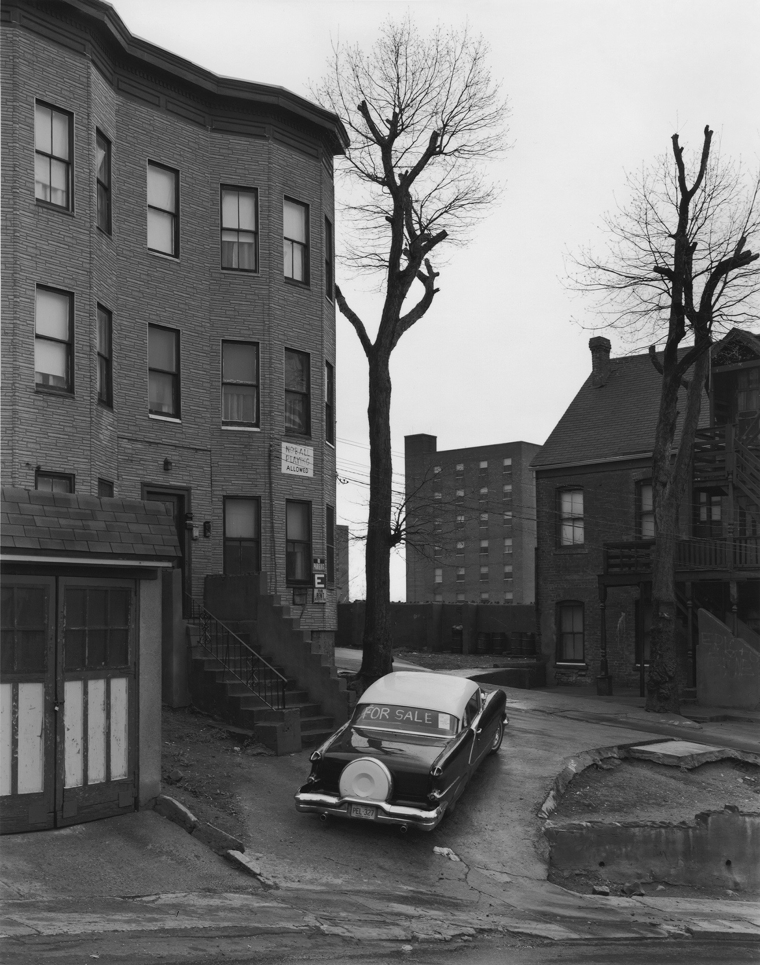 Photographie noir et blanc d'immeuble et de voiture, dans une banlieue américaine des années 1960-1970.